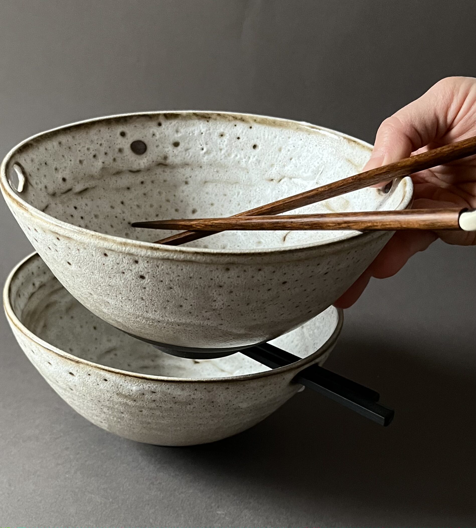 Deux bols à ramen Tommy empilés, tenus en main, baguettes en bois et noires glissées dans les trous