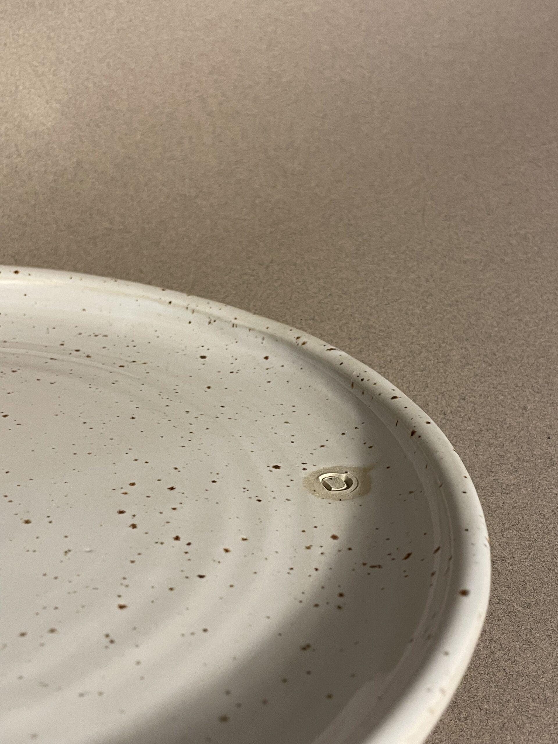 Gros plan sur le rebord de l'assiette plate Maya, émail blanc moucheté et logo adb en creux