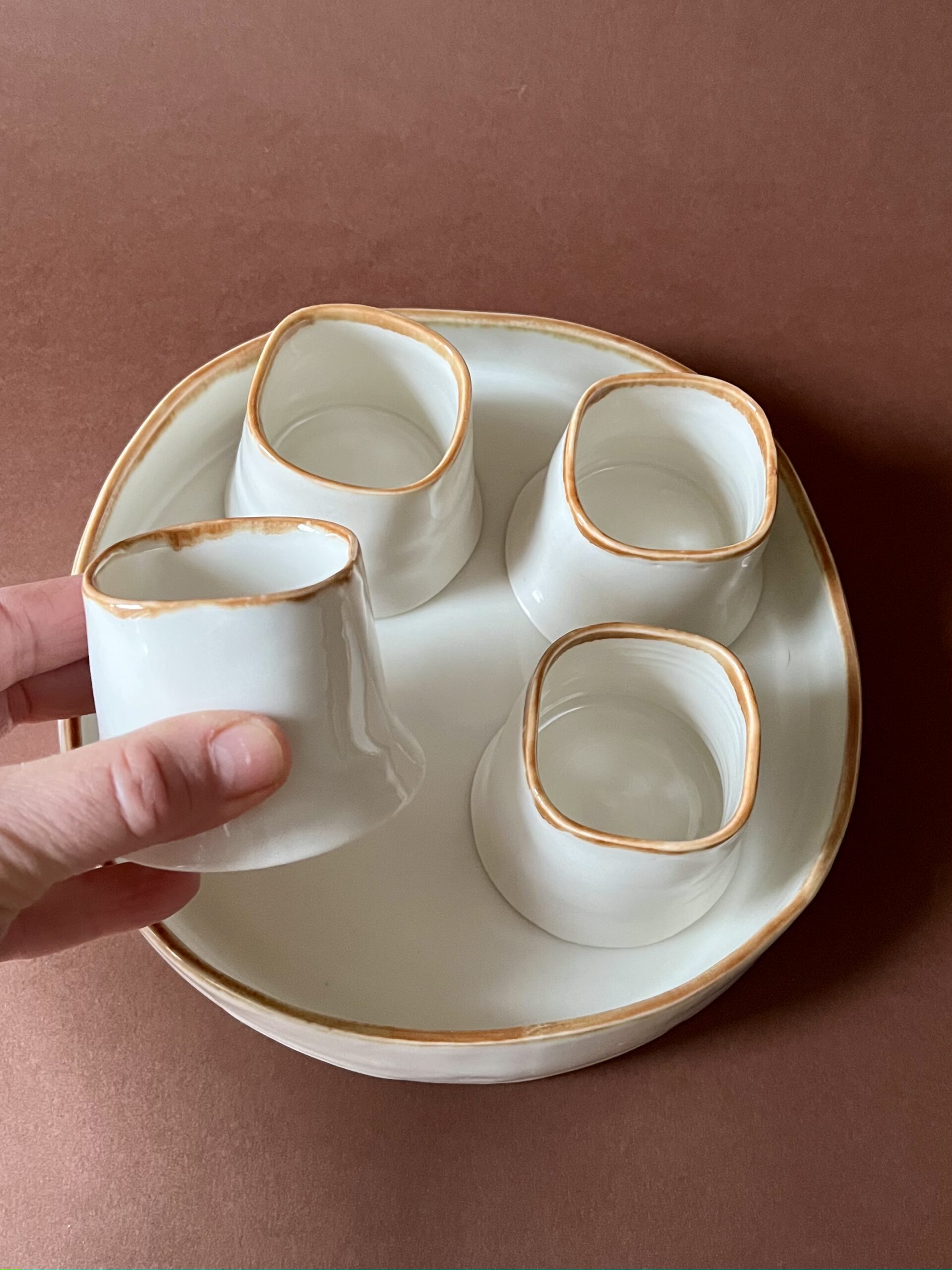 Tasse Ulysse à liseré marron tenue en main au-dessus du plateau et des trois autres tasses