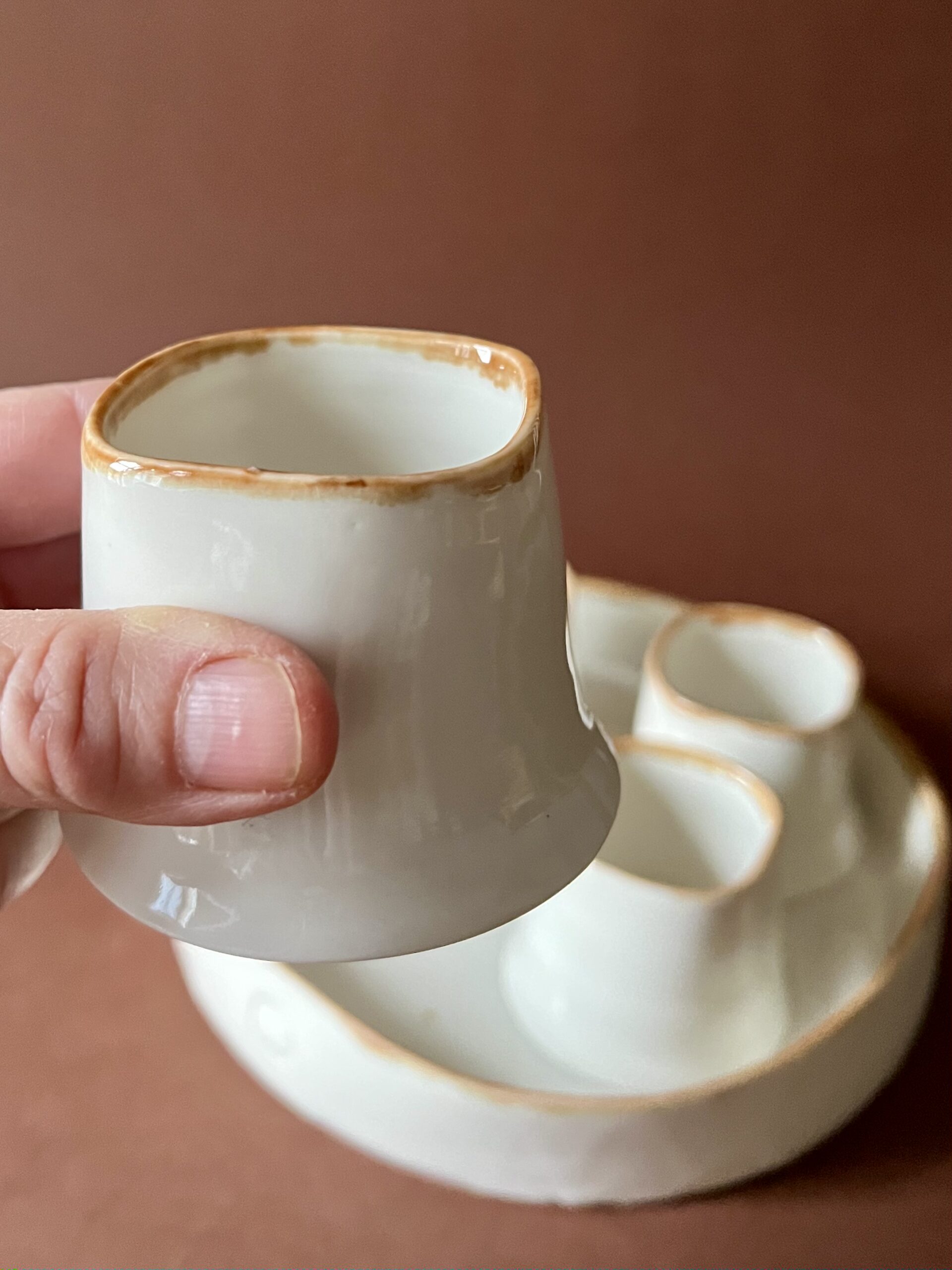 Tasse Ulysse à liseré marron tenue en main montrant sa forme carrée concave, plateau et tasses derrière