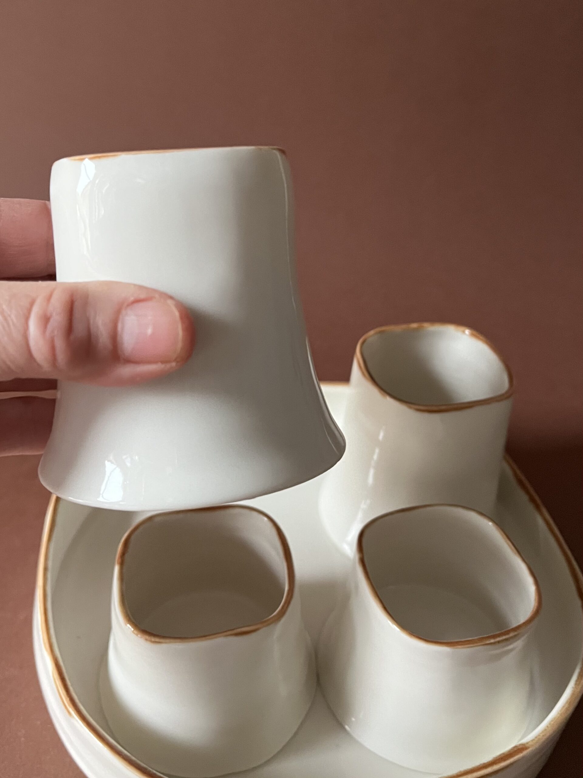 Tasse Ulysse à liseré marron tenue en main, dessous en porcelaine visible, autres tasses sur le plateau