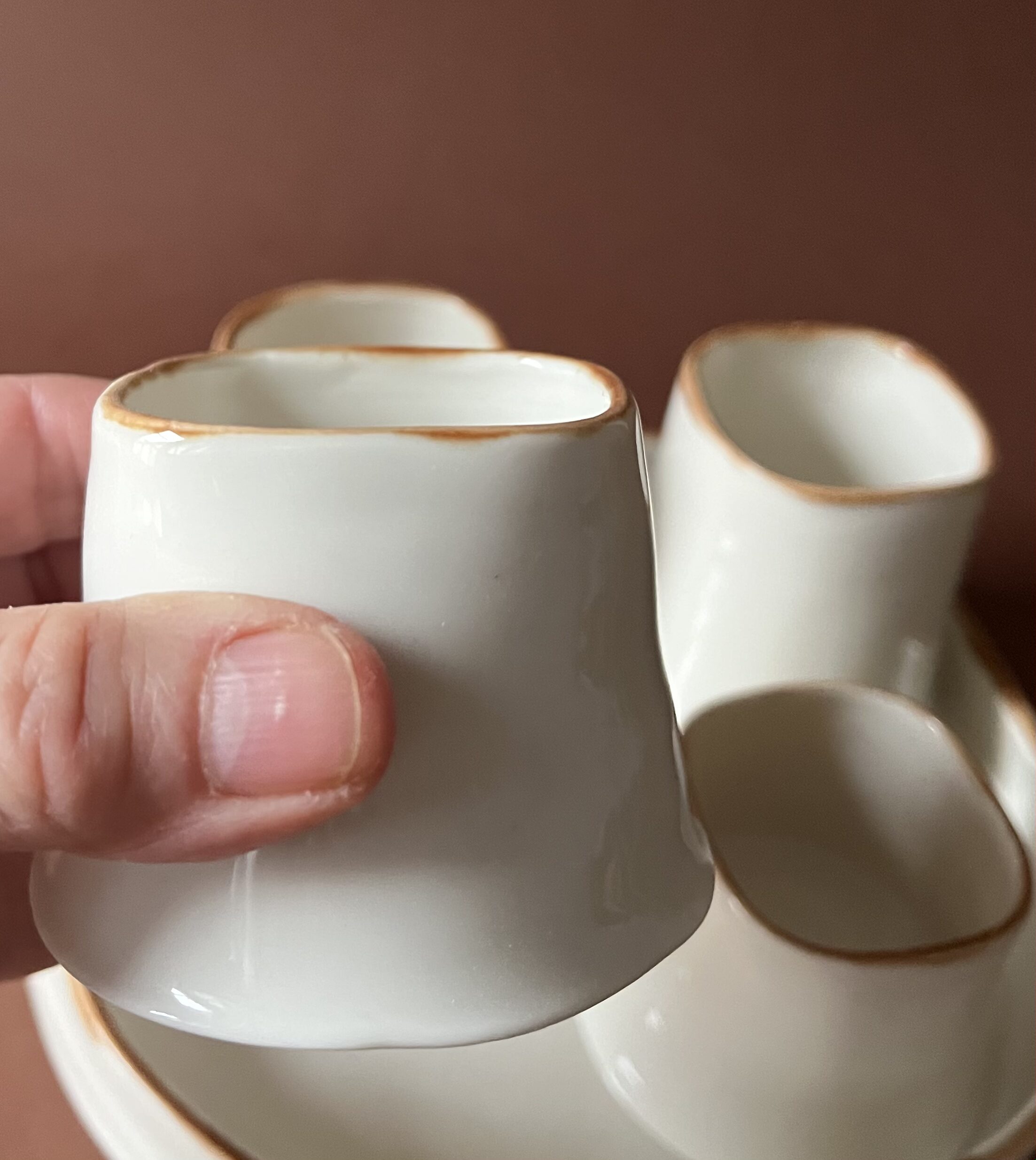 Tasse Ulysse à liseré marron tenue en main montrant son ouverture carrée, autres tasses derrière