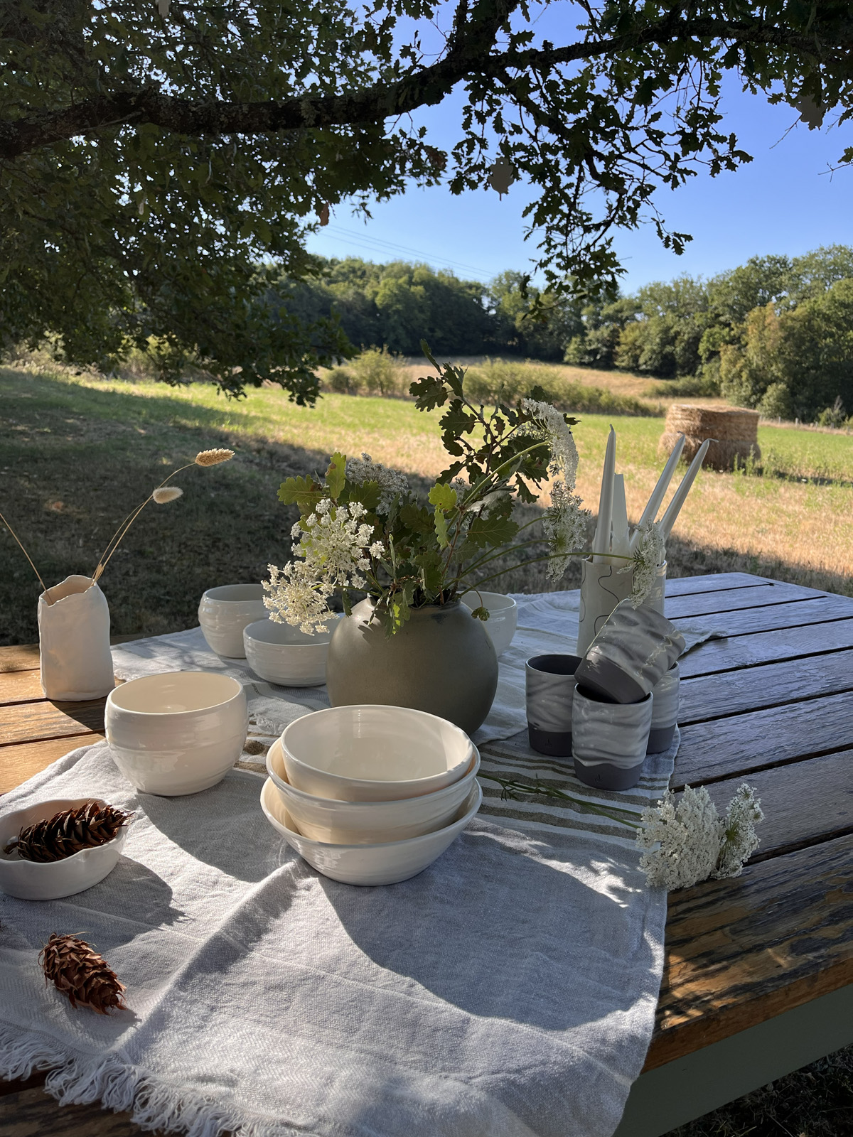 Table de jardin en bois dressée de bols, gobelets et vases en grès sous un arbre, champ en arrière-plan