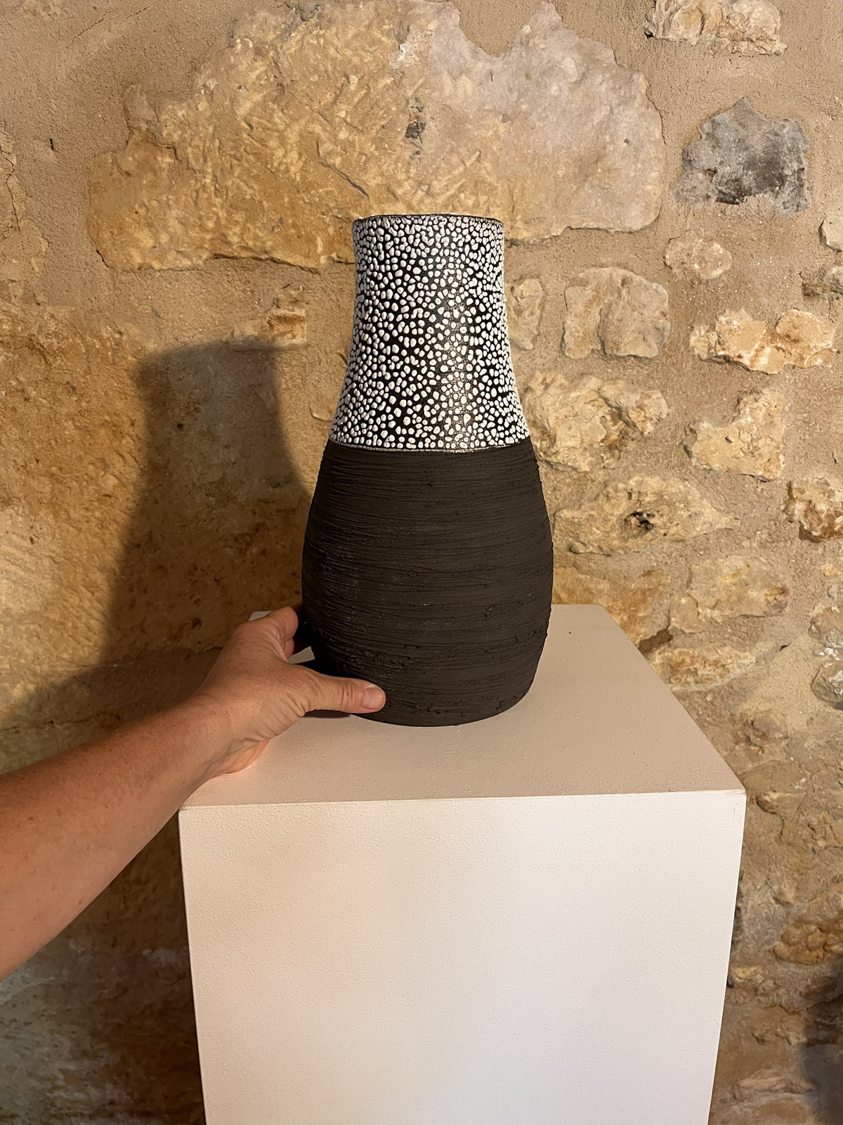 Main posant un vase Winter en grès noir strié à col émaillé blanc sur un socle, mur de pierre