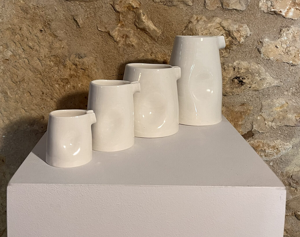 Quatre saucières verseuses en porcelaine blanche de tailles croissantes alignées sur un socle, mur de pierre