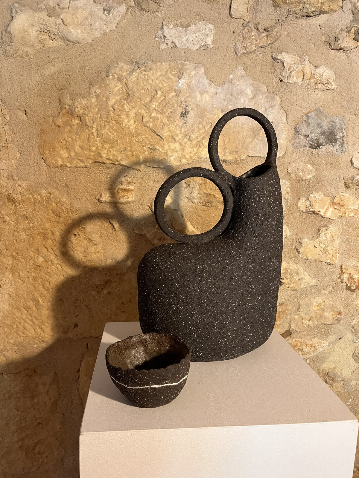 Vase sculptural en grès noir à deux anneaux et petit bol noir à filet blanc, sur socle devant un mur de pierre