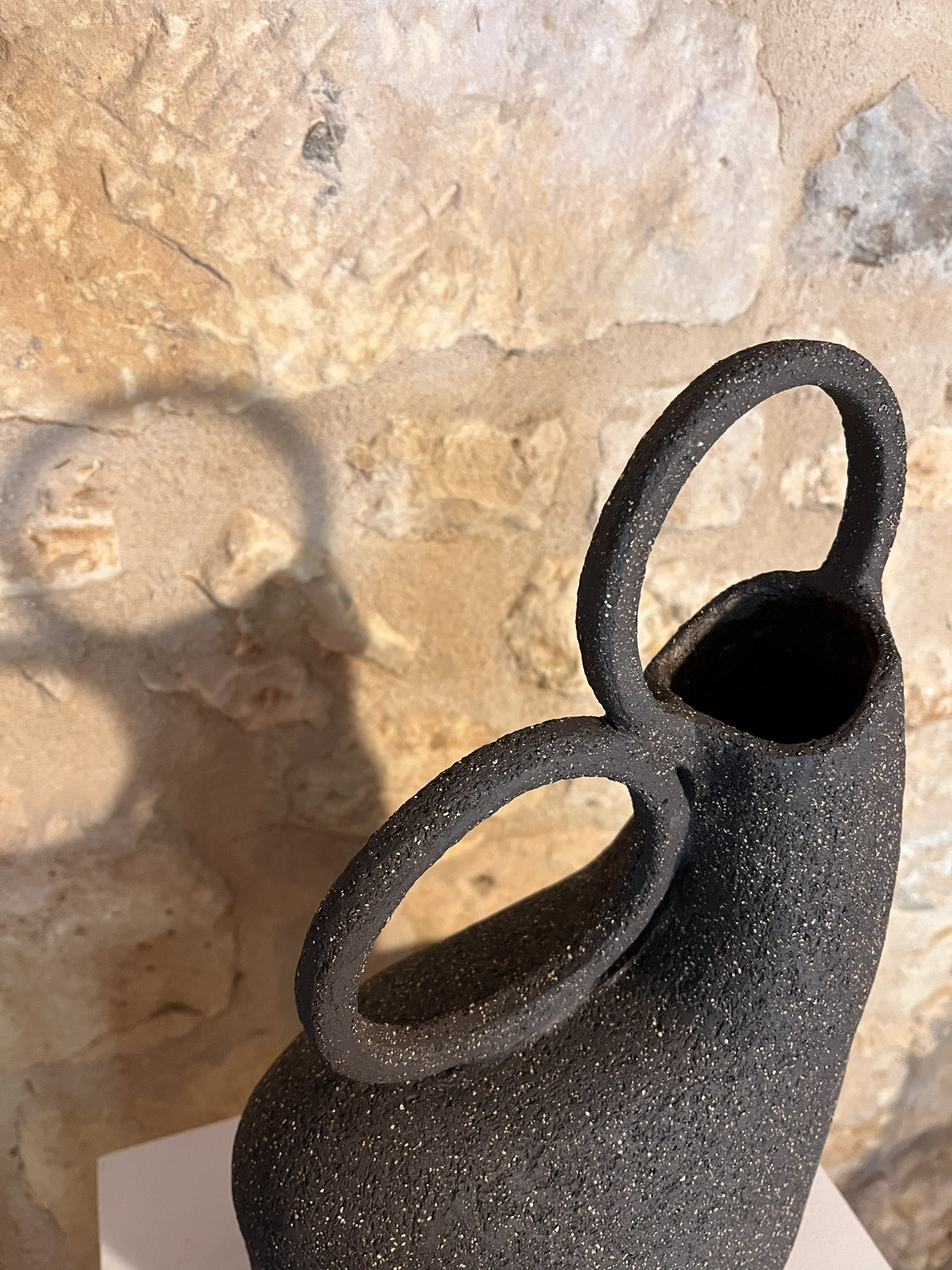 Gros plan sur l'ouverture et les deux anneaux d'un vase sculptural en grès noir chamotté, ombre sur le mur