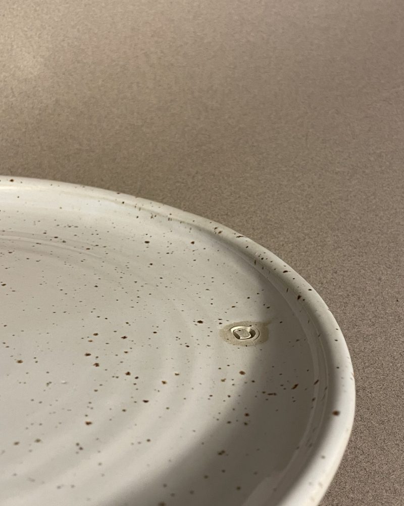 Gros plan sur le rebord de l'assiette plate Maya, émail blanc moucheté et logo adb en creux