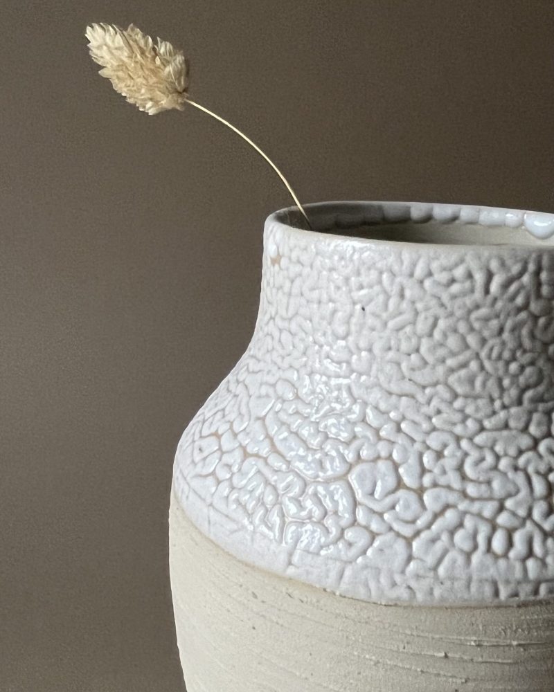 Gros plan sur le col large émaillé blanc rétracté d'un vase Winter en grès beige strié, graminée