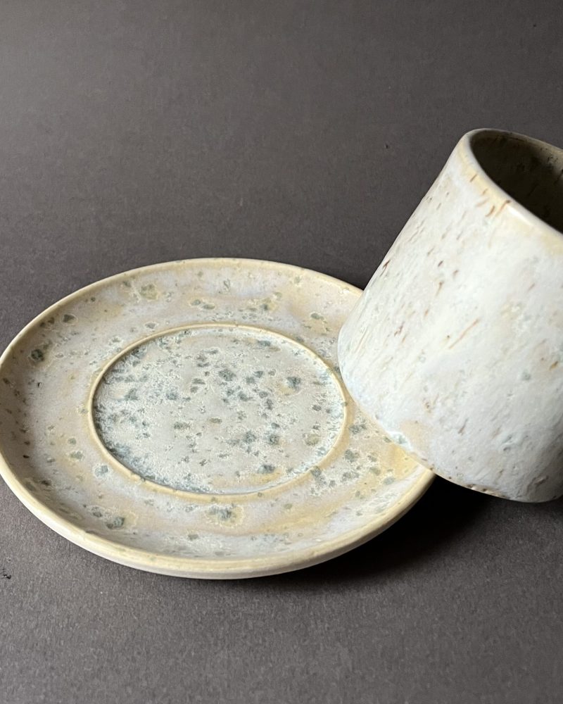 Tasse Steph en grès à l'émail satiné beige et vert appuyée sur le bord de sa soucoupe assortie