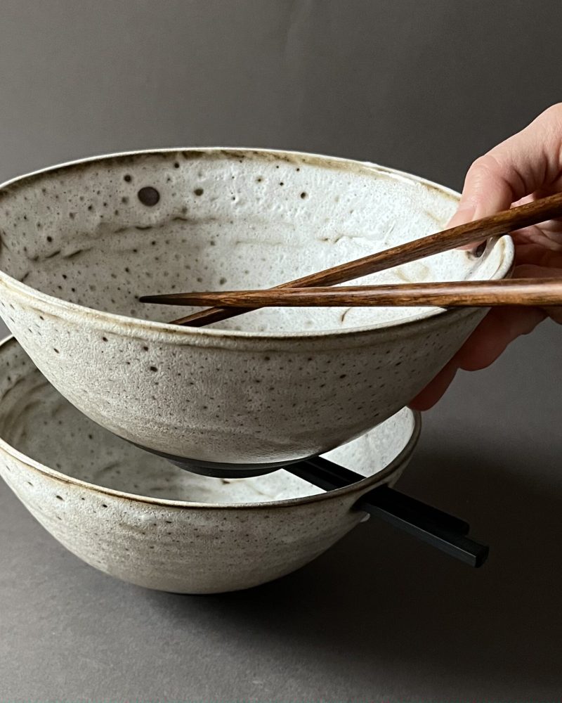 Deux bols à ramen Tommy empilés, tenus en main, baguettes en bois et noires glissées dans les trous