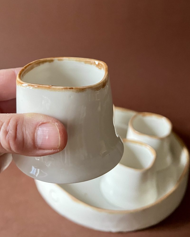 Tasse Ulysse à liseré marron tenue en main montrant sa forme carrée concave, plateau et tasses derrière