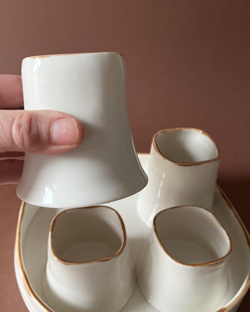 Tasse Ulysse à liseré marron tenue en main, dessous en porcelaine visible, autres tasses sur le plateau