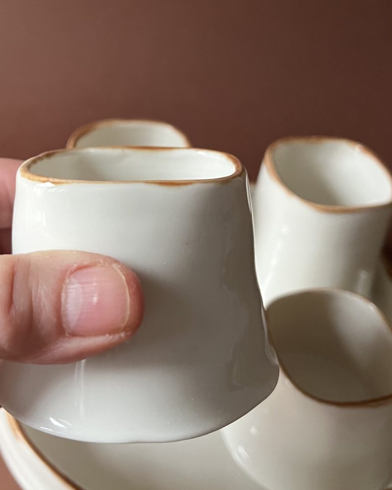 Tasse Ulysse à liseré marron tenue en main montrant son ouverture carrée, autres tasses derrière