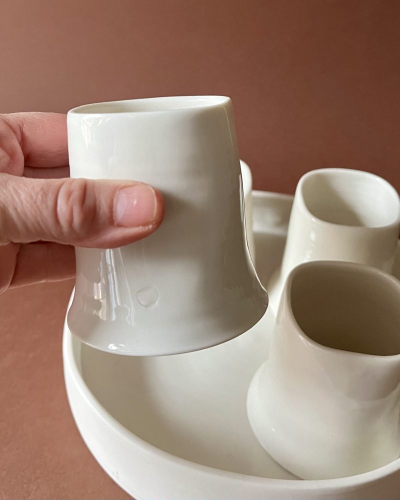 Tasse Ulysse blanche tenue en main au-dessus du set, tasses et plateau en porcelaine blanche