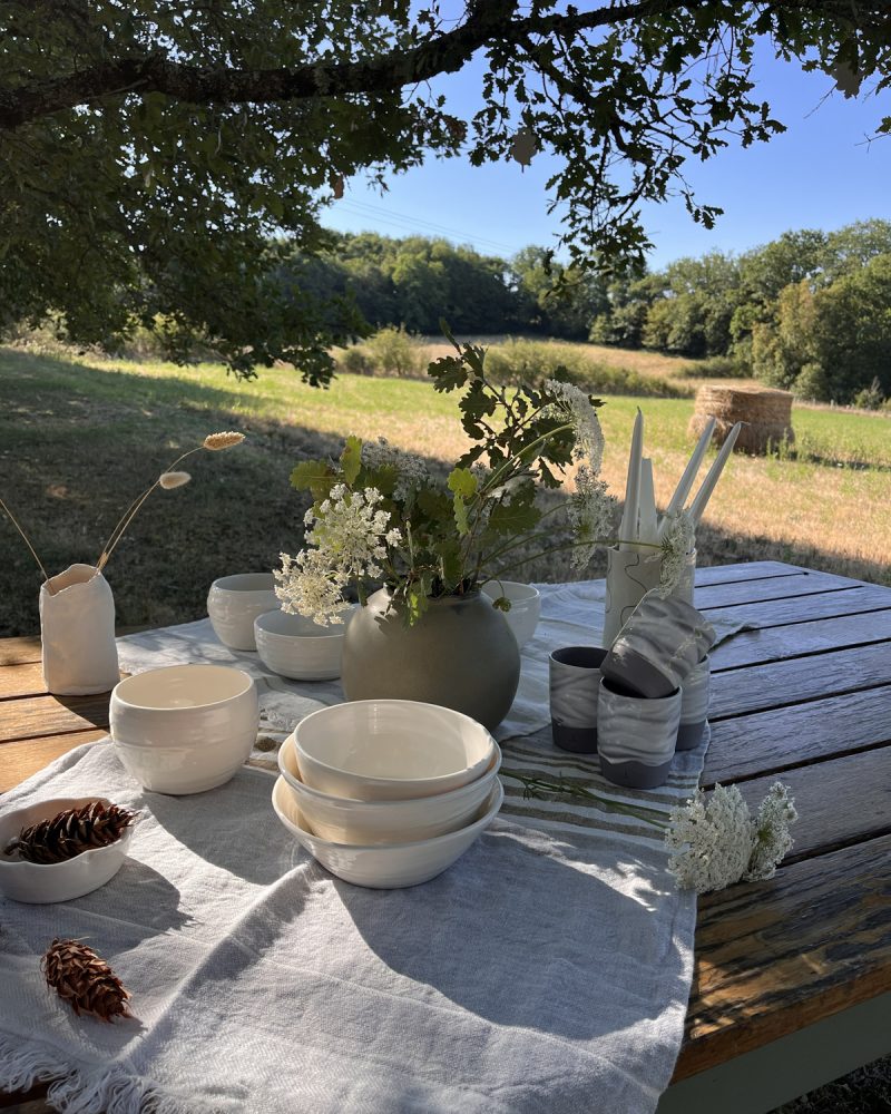 Table de jardin en bois dressée de bols, gobelets et vases en grès sous un arbre, champ en arrière-plan