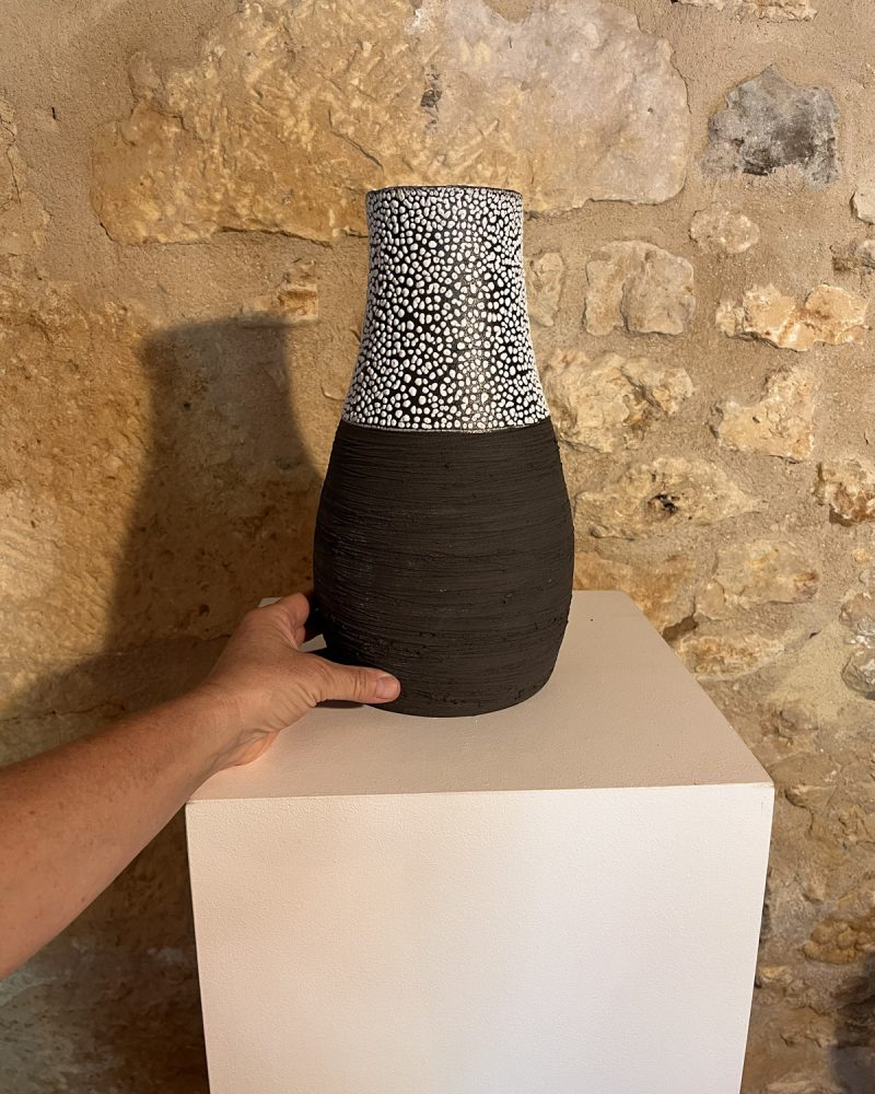 Main posant un vase Winter en grès noir strié à col émaillé blanc sur un socle, mur de pierre