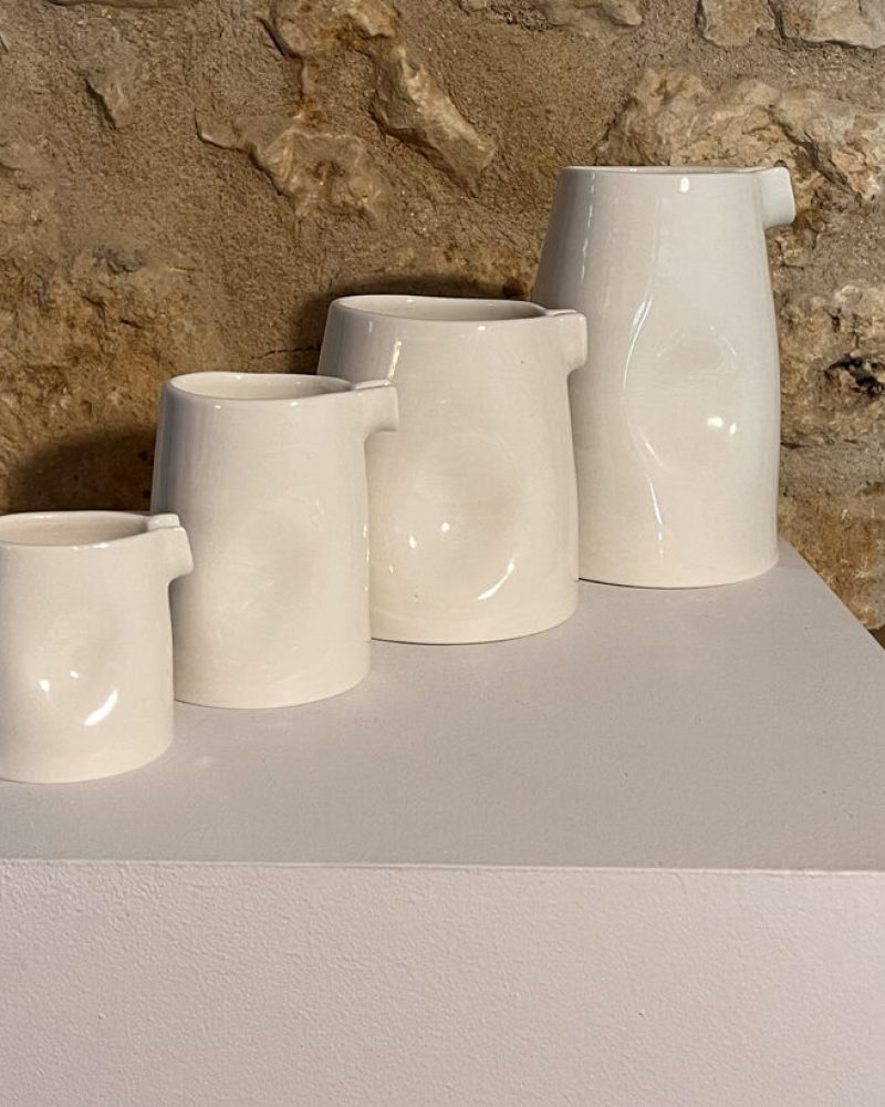 Quatre saucières verseuses en porcelaine blanche de tailles croissantes alignées sur un socle, mur de pierre