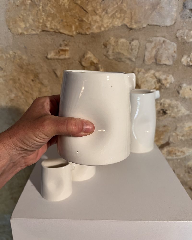 Main tenant la grande saucière verseuse en porcelaine blanche par son renfoncement, petites saucières derrière