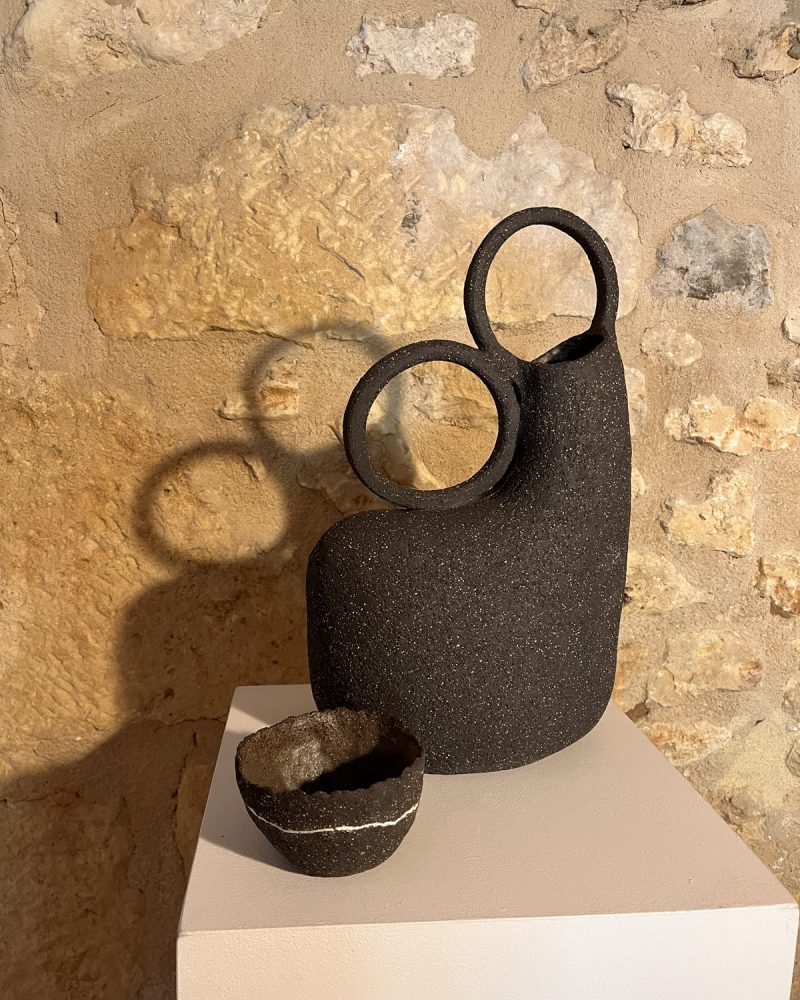 Vase sculptural en grès noir à deux anneaux et petit bol noir à filet blanc, sur socle devant un mur de pierre