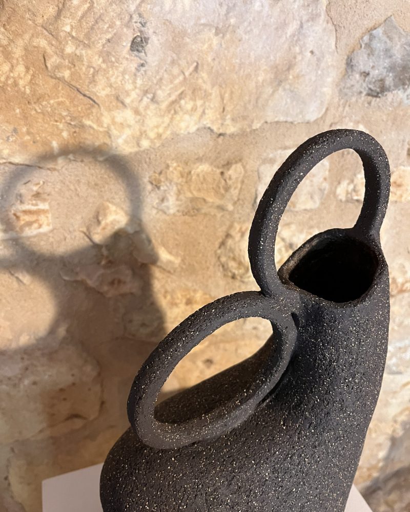 Gros plan sur l'ouverture et les deux anneaux d'un vase sculptural en grès noir chamotté, ombre sur le mur
