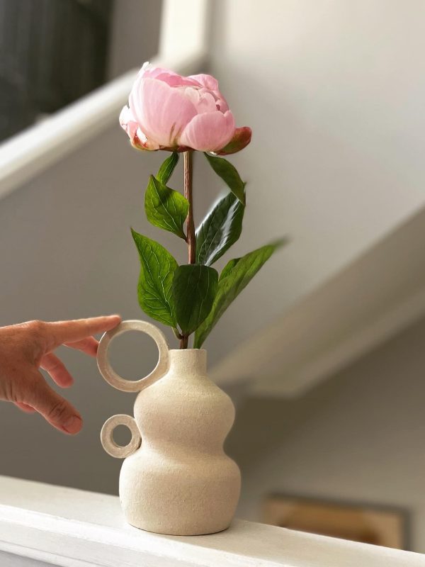 Soliflore en grès beige à anneaux garni d'une pivoine rose sur une console, main tenant la fleur