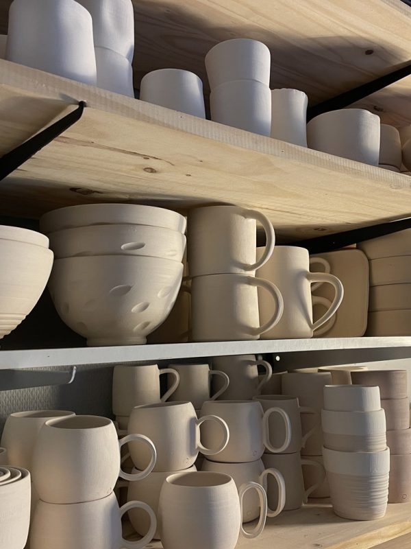 Étagères en bois de l'atelier chargées de bols, tasses et assiettes en biscuit blanc