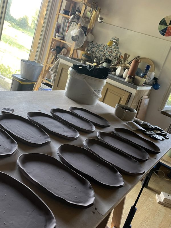Rangées d'assiettes ovales en terre noire crue posées sur une table de l'atelier au soleil