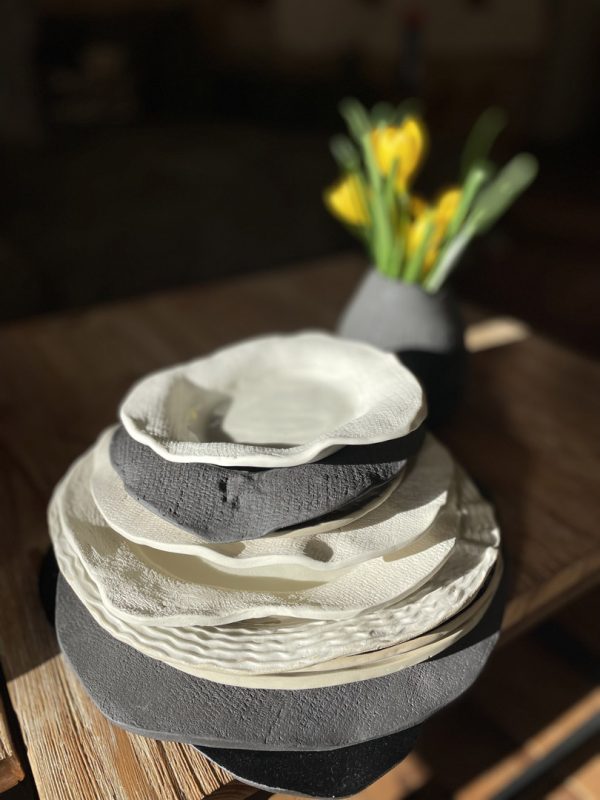 Pile d'assiettes en grès blanc et noir aux bords irréguliers et textures variées, tulipes jaunes en fond